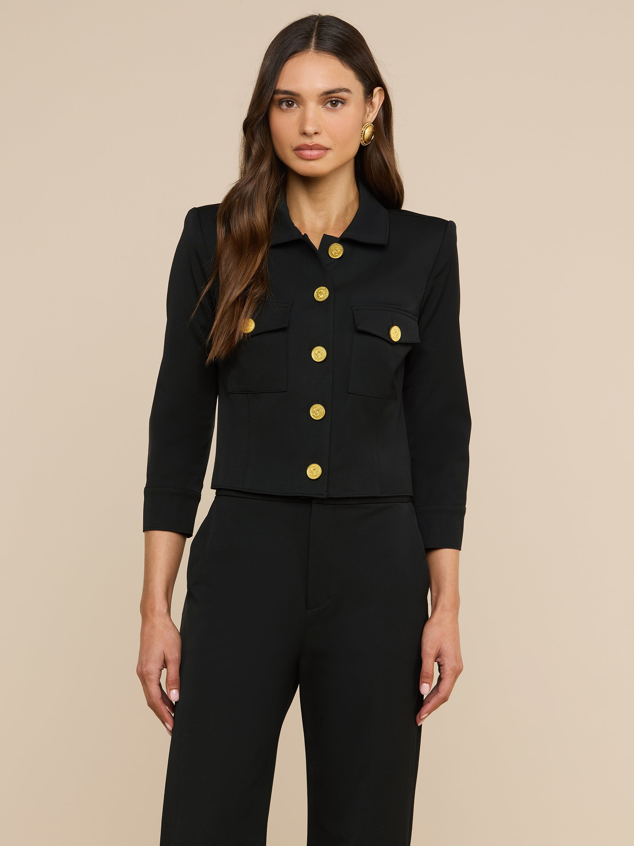 Eine Frau mit langen, welligen braunen Haaren trägt die „Kumi Cropped Jacket“ – eine schwarze, taillierte Jacke mit Dreiviertelärmeln, goldenen Knöpfen und Pattentaschen –, dazu eine passende schwarze Hose, und steht vor einem schlichten beigen Hintergrund.