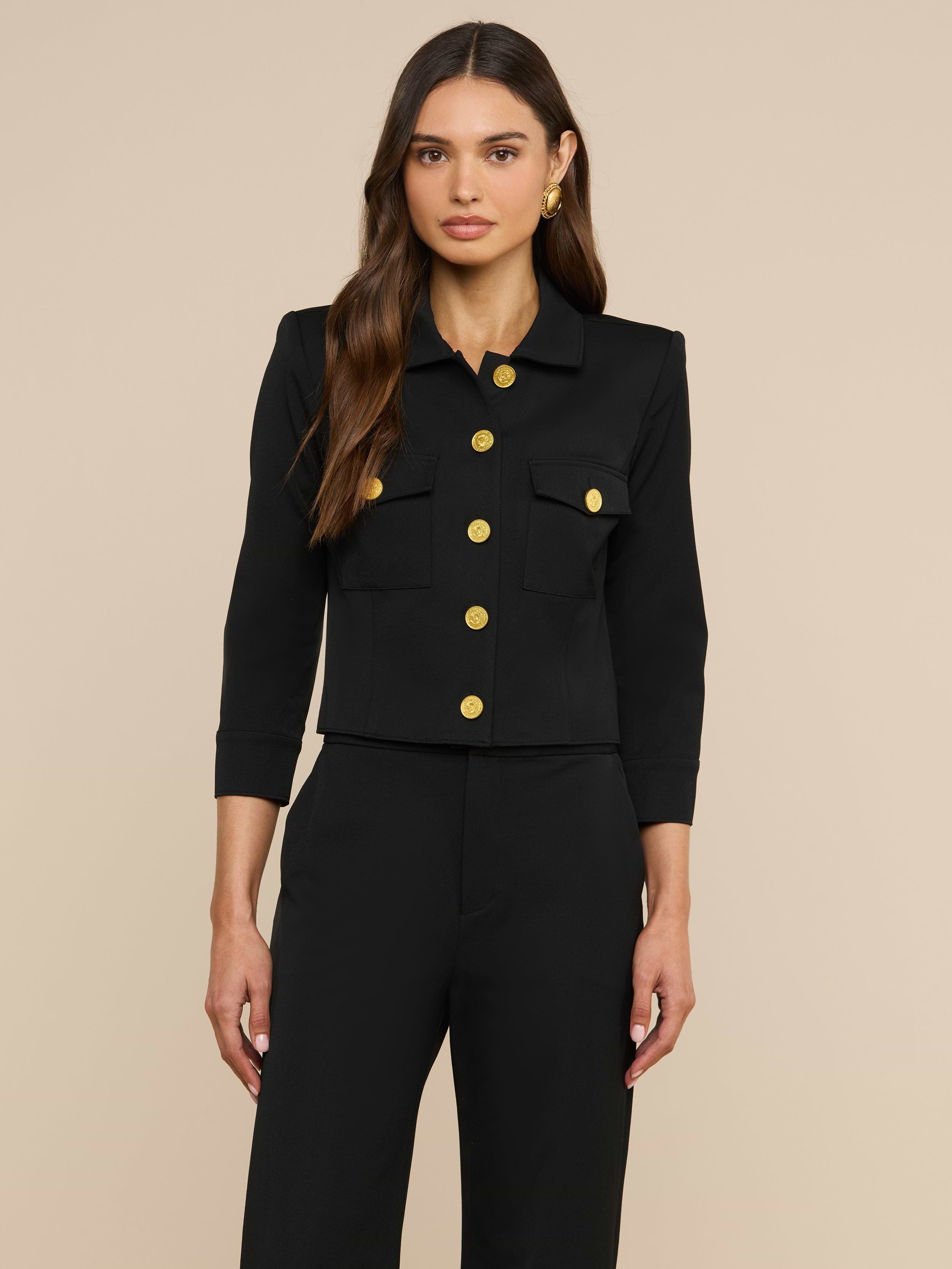Eine Frau mit langen, welligen braunen Haaren trägt die „Kumi Cropped Jacket“ – eine schwarze, taillierte Jacke mit Dreiviertelärmeln, goldenen Knöpfen und Pattentaschen –, dazu eine passende schwarze Hose, und steht vor einem schlichten beigen Hintergrund.