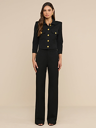 Eine Frau steht mit dem Blick nach vorne vor einem beigen Hintergrund und trägt die „Kumi Cropped Jacket“ mit goldenen Knöpfen sowie eine Hose mit weitem Bein aus elastischem Anzugstoff.