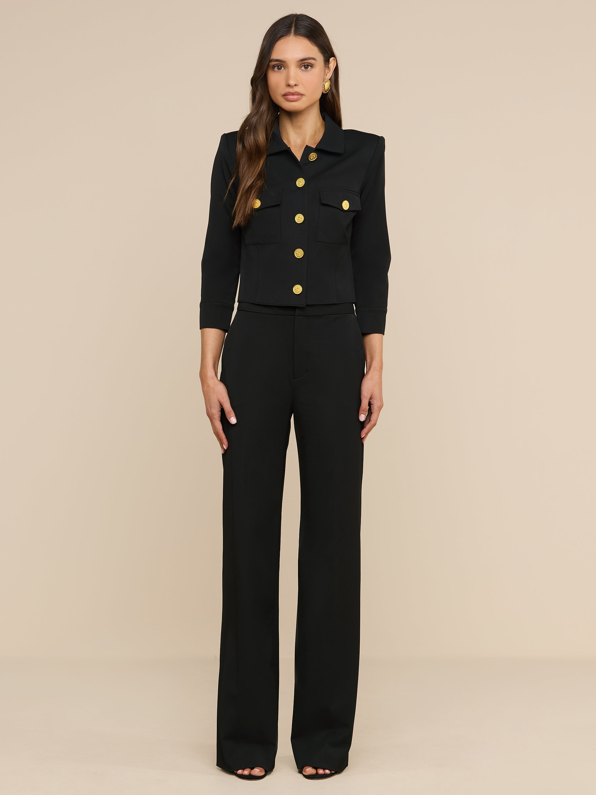 Eine Frau steht mit dem Blick nach vorne vor einem beigen Hintergrund und trägt die „Kumi Cropped Jacket“ mit goldenen Knöpfen sowie eine Hose mit weitem Bein aus elastischem Anzugstoff.