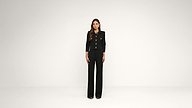 A woman stands facing forward in a minimalist studio, wearing a black pantsuit with gold buttons. The background is plain and white.