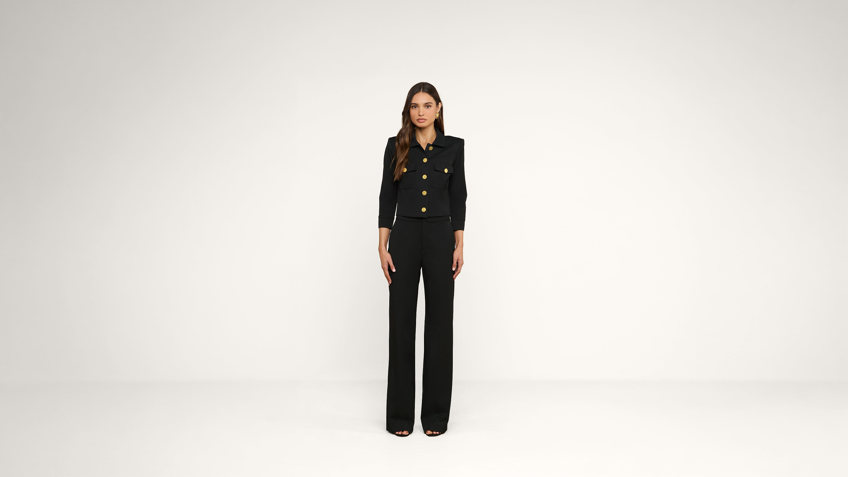 A woman stands facing forward in a minimalist studio, wearing a black pantsuit with gold buttons. The background is plain and white.