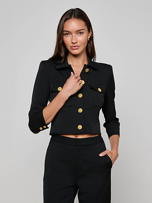 A woman wearing the L'AGENCE Kumi Cropped Jacket with gold buttons and matching pants stands against a plain light background. She has long brown hair, gold earrings, and poses with one hand near her collar and the other in her pocket.