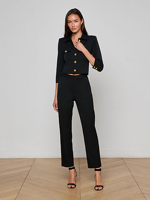 A woman stands against a white wall, wearing the L'AGENCE Kumi Cropped Jacket—black stretch suit fabric with gold buttons and three-quarter sleeves—paired with high-waisted black pants, black heels, and her long hair loose.