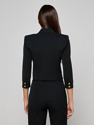 A woman with long brown hair is seen from behind wearing the L'AGENCE Kumi Cropped Jacket, a fitted black piece with three-quarter sleeves and gold buttons, crafted from stretch suit fabric, paired with matching pants against a light background.