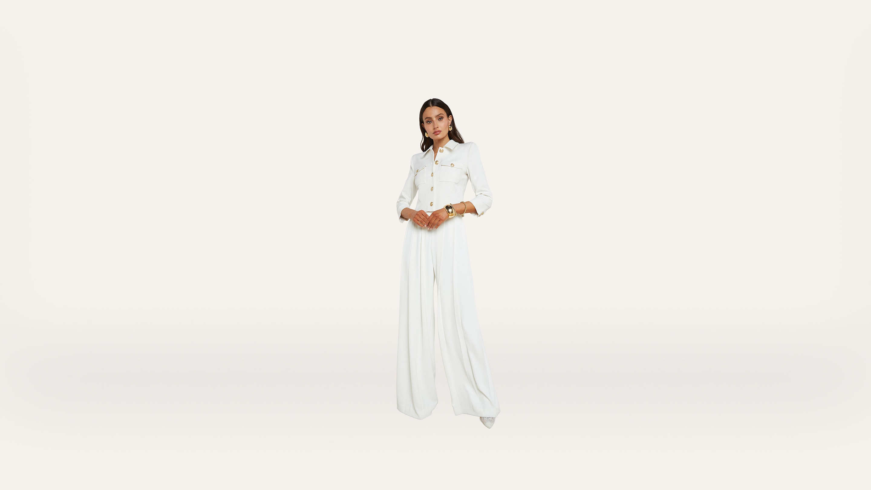 A person stands centered against a plain white background, wearing a long-sleeved white button-up top and flowing white pants, with hands clasped at the waist.