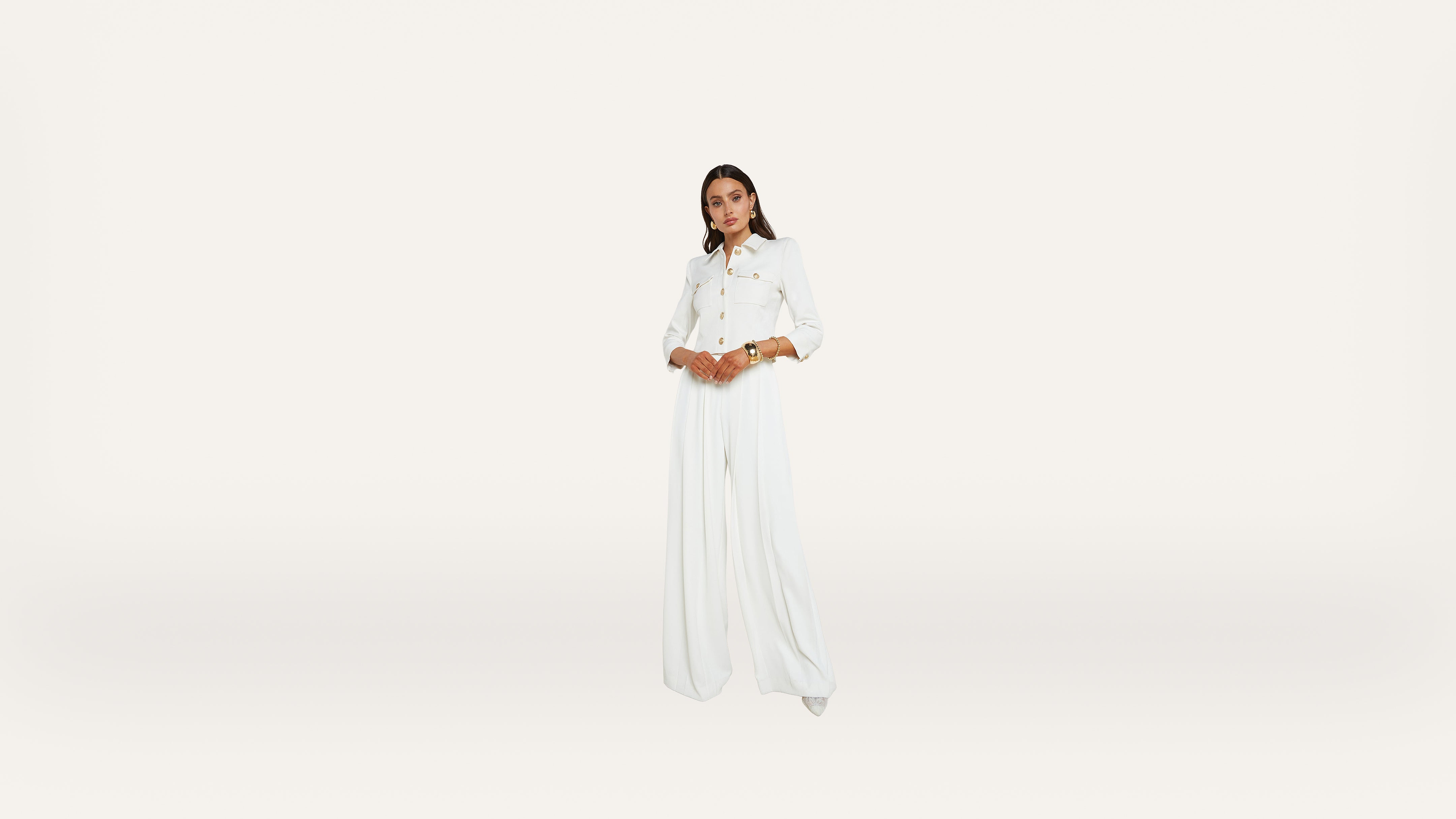 A person stands centered against a plain white background, wearing a long-sleeved white button-up top and flowing white pants, with hands clasped at the waist.