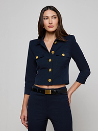 A woman wearing the Kumi Cropped Jacket in midnight navy, featuring gold buttons and three-quarter sleeves, stands against a plain light background. The jacket, crafted from stretch suit fabric for comfort and style, is paired with matching navy pants as she looks slightly to her left with her left hand resting near her pocket.