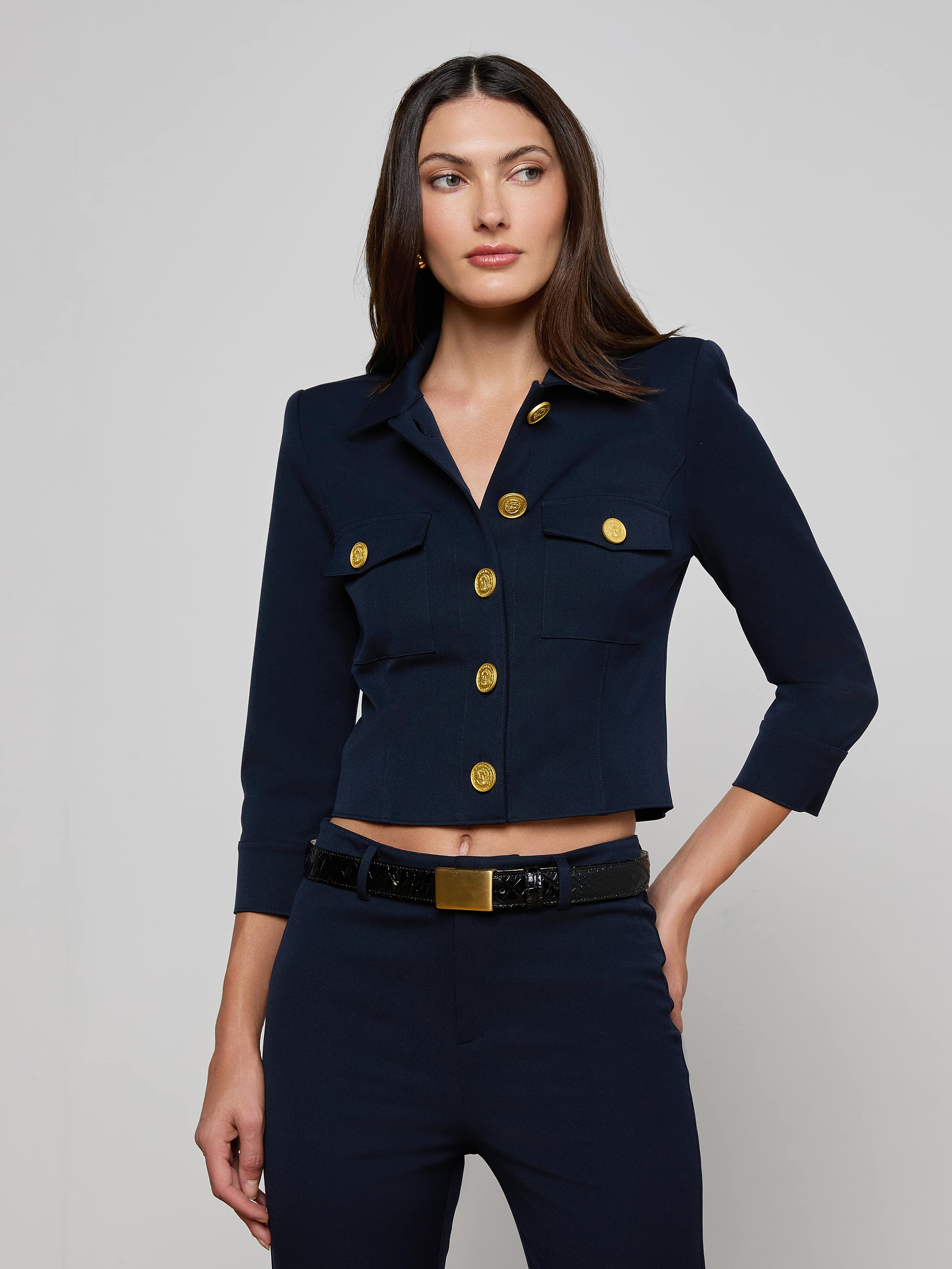 A woman wearing the Kumi Cropped Jacket in midnight navy, featuring gold buttons and three-quarter sleeves, stands against a plain light background. The jacket, crafted from stretch suit fabric for comfort and style, is paired with matching navy pants as she looks slightly to her left with her left hand resting near her pocket.
