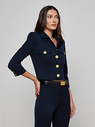 A woman wearing the Kumi Cropped Jacket—crafted from stretch suit fabric in navy, featuring gold buttons and chest pockets, styled with a black belt with a gold buckle—stands against a plain light background.