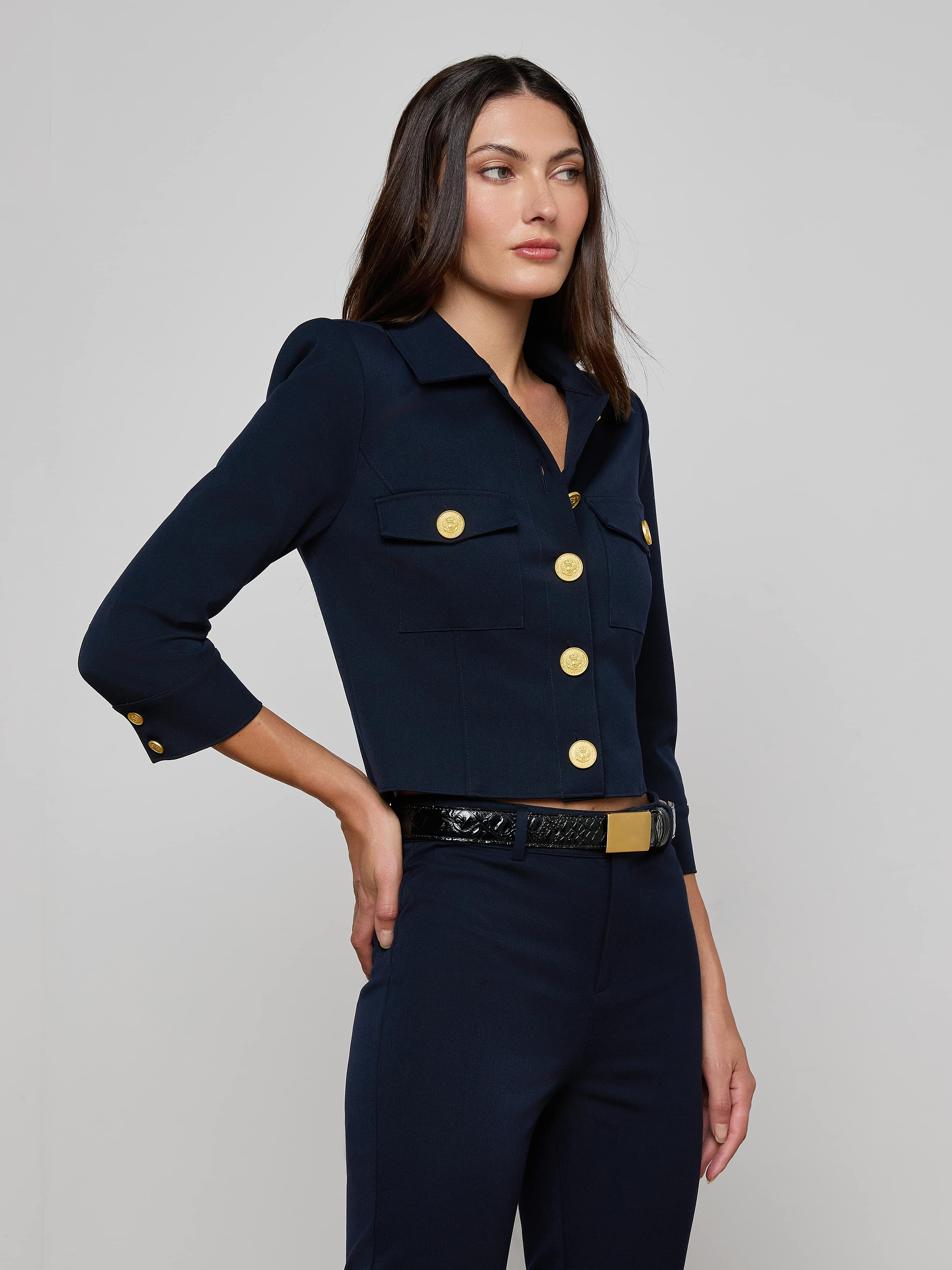 A woman wearing the Kumi Cropped Jacket—crafted from stretch suit fabric in navy, featuring gold buttons and chest pockets, styled with a black belt with a gold buckle—stands against a plain light background.