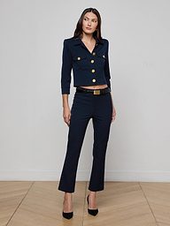 A woman stands against a plain white background wearing the Kumi Cropped Jacket in midnight navy, featuring gold buttons and a matching belt. She pairs it with black high heels and maintains a neutral expression.