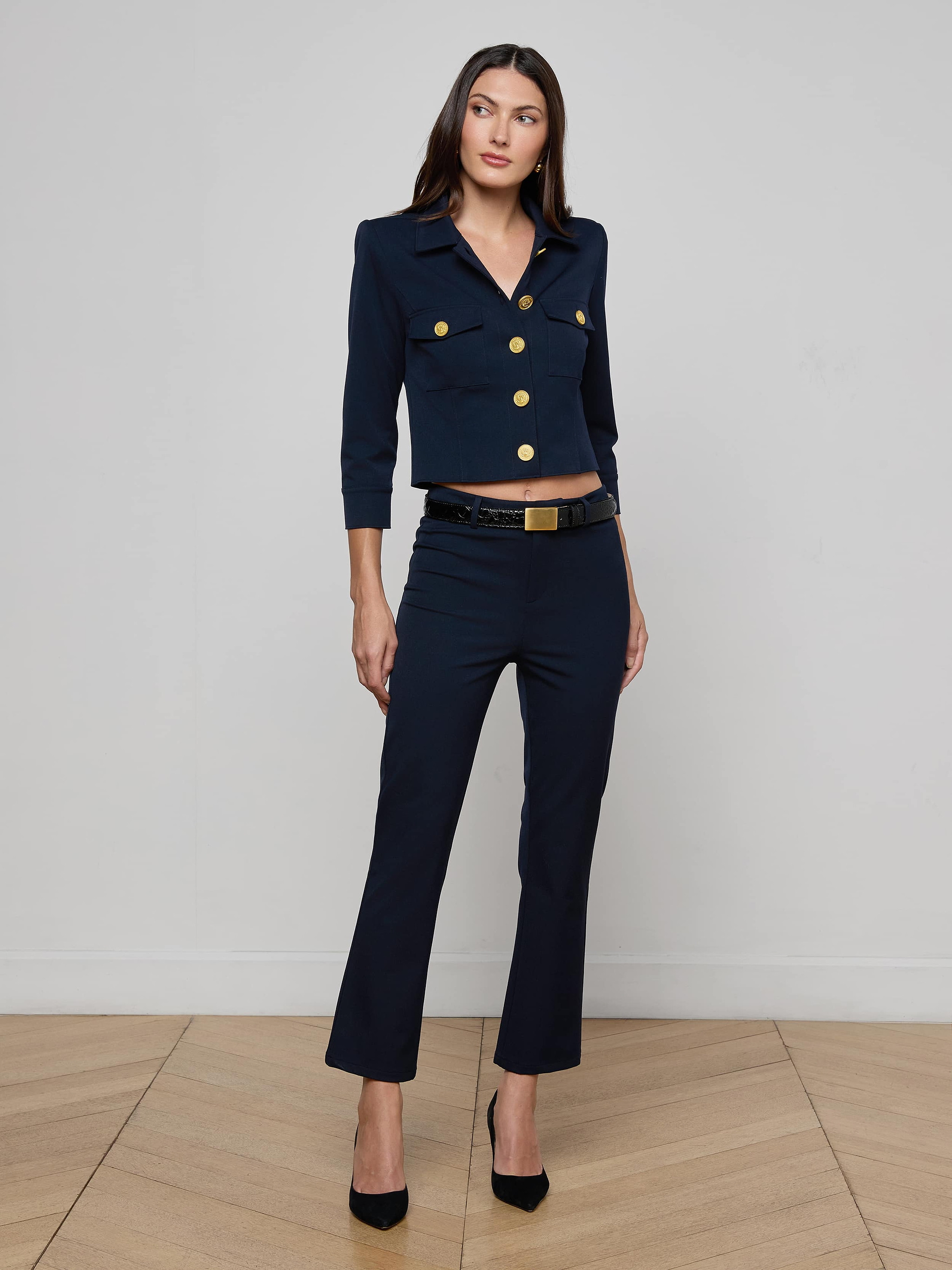 A woman stands against a plain white background wearing the Kumi Cropped Jacket in midnight navy, featuring gold buttons and a matching belt. She pairs it with black high heels and maintains a neutral expression.