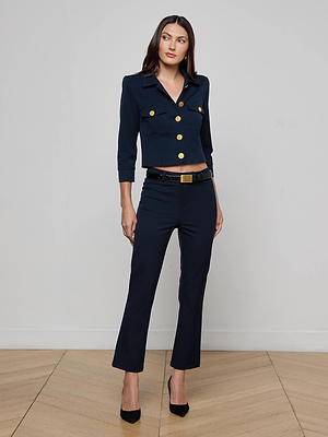 A woman with long straight hair and a neutral expression stands indoors on a wooden floor, wearing the L'AGENCE Kumi Cropped Jacket in midnight navy stretch fabric, paired with matching pants, a black belt, and black pointed-toe heels.