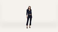 A woman wearing a navy suit and black heels stands against a plain white background, looking slightly to her right with a relaxed posture.