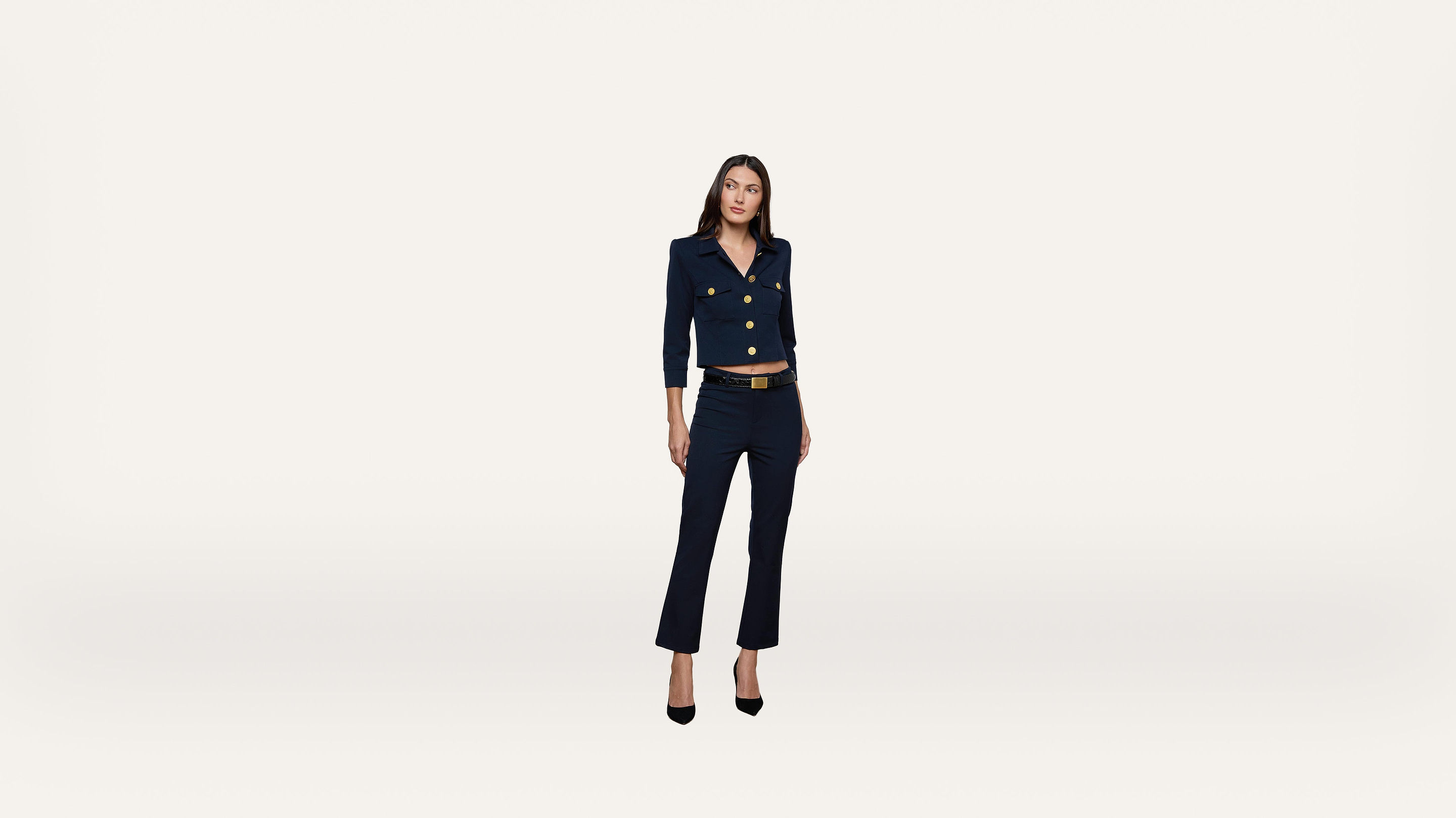 A woman wearing a navy suit and black heels stands against a plain white background, looking slightly to her right with a relaxed posture.