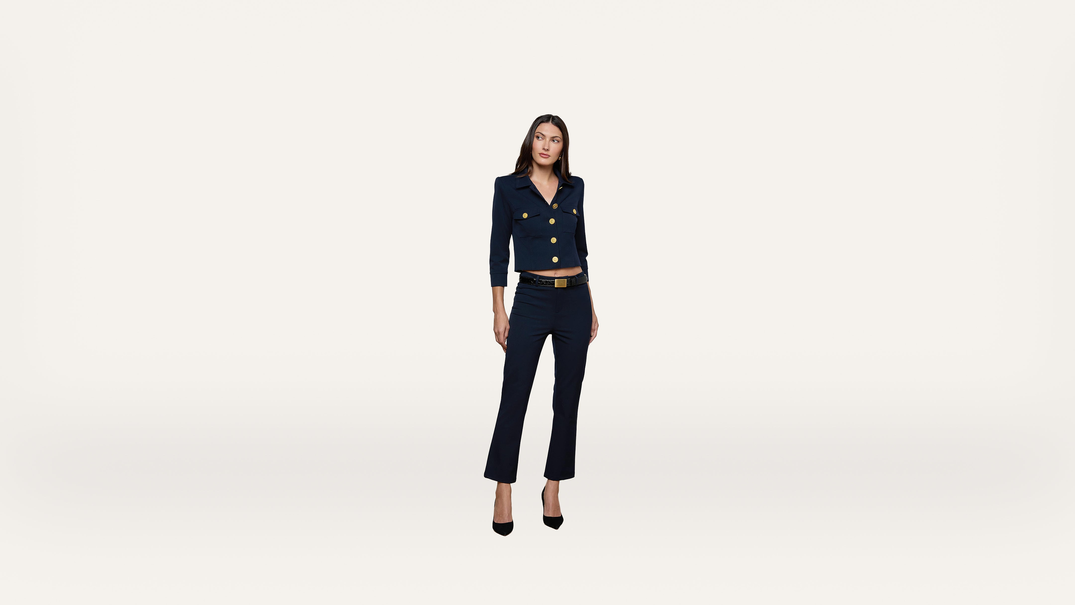 A woman wearing a navy suit and black heels stands against a plain white background, looking slightly to her right with a relaxed posture.