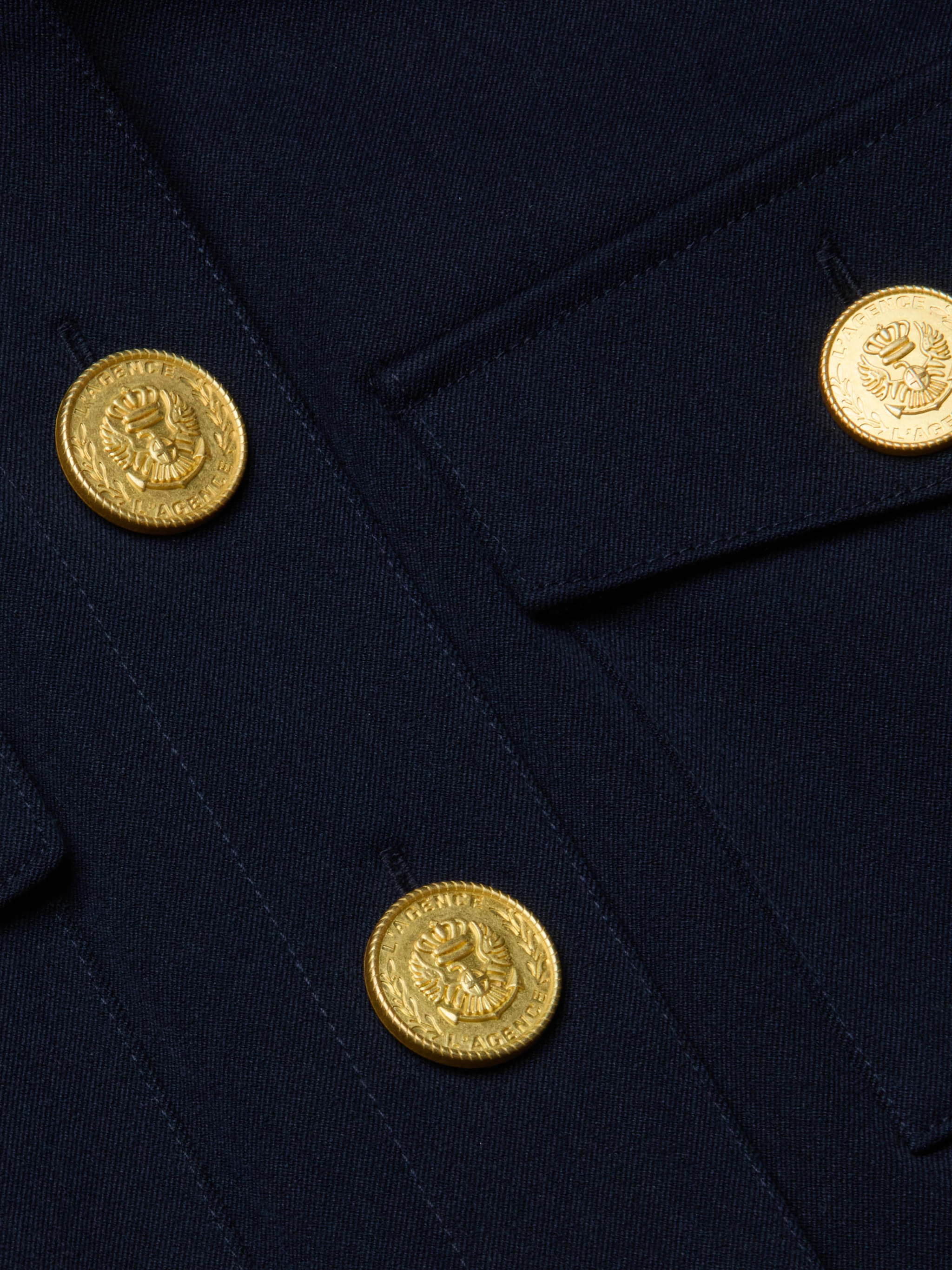 Close-up of the Kumi Cropped Jacket in midnight navy stretch suit fabric, showcasing gold lion head-embossed decorative buttons beside a flap pocket and seam—evoking a formal, military-inspired look with three-quarter sleeves.