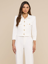 A woman stands facing forward, wearing the Kumi Cropped Jacket in white stretch-suiting fabrication with gold embossed dome buttons, paired with matching white pants, against a plain light background. She has long brown hair and a neutral expression.