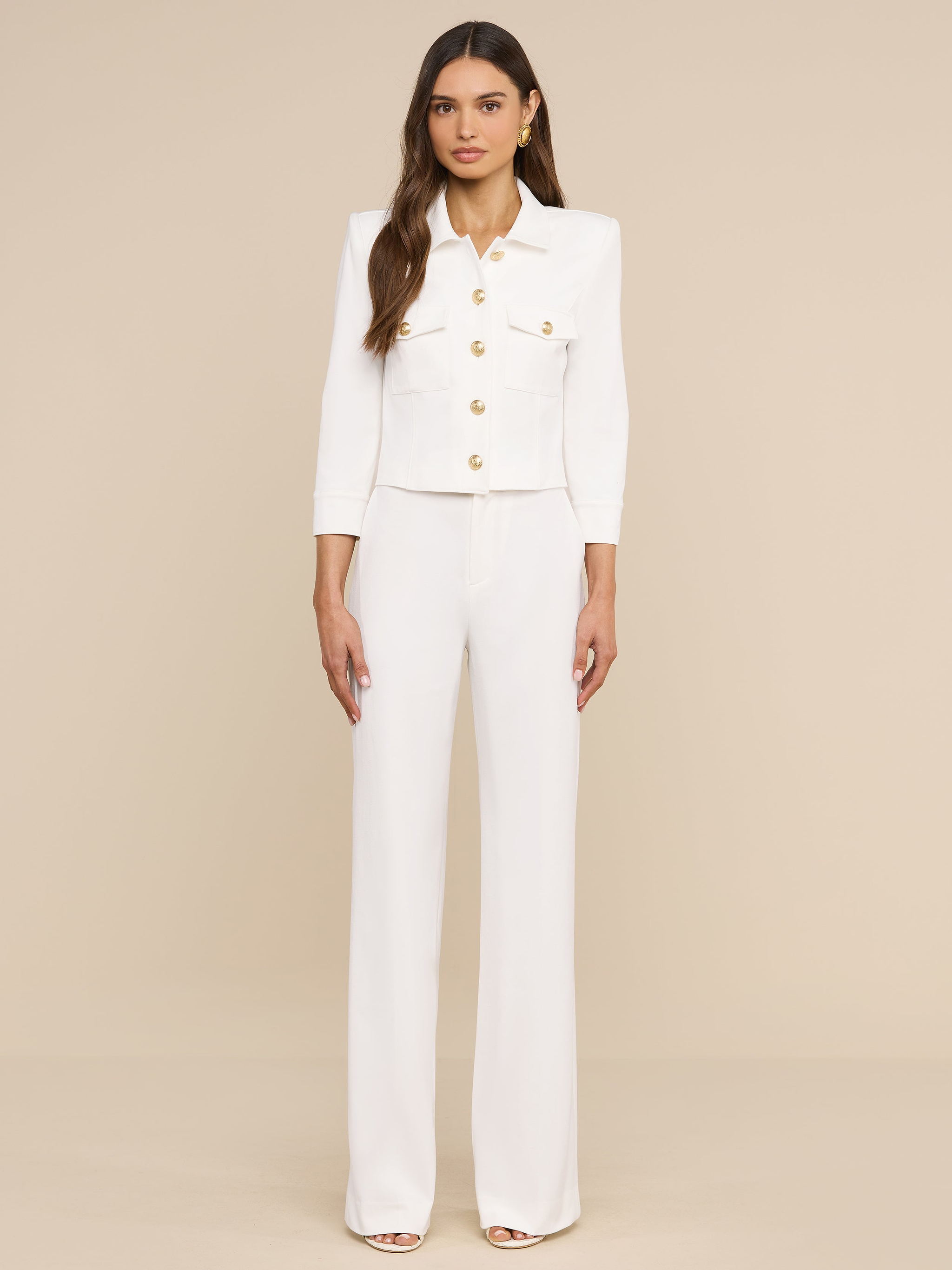 A woman stands against a plain white background, wearing the Kumi Cropped Jacket crafted from stretch suiting with three-quarter sleeves and gold-embossed dome buttons, paired with matching wide-leg white pants.