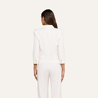 A woman with long brown hair stands facing away, wearing the Kumi Cropped Jacket in white stretch-suiting fabrication with gold embossed dome buttons and matching white pants against a plain light background.