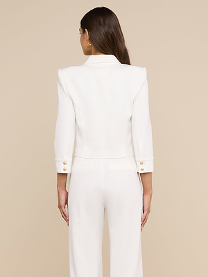 Una mujer con el pelo largo y castaño, que lleva la chaqueta corta «Kumi» en color blanco, está de espaldas a la cámara frente a un fondo beige.
