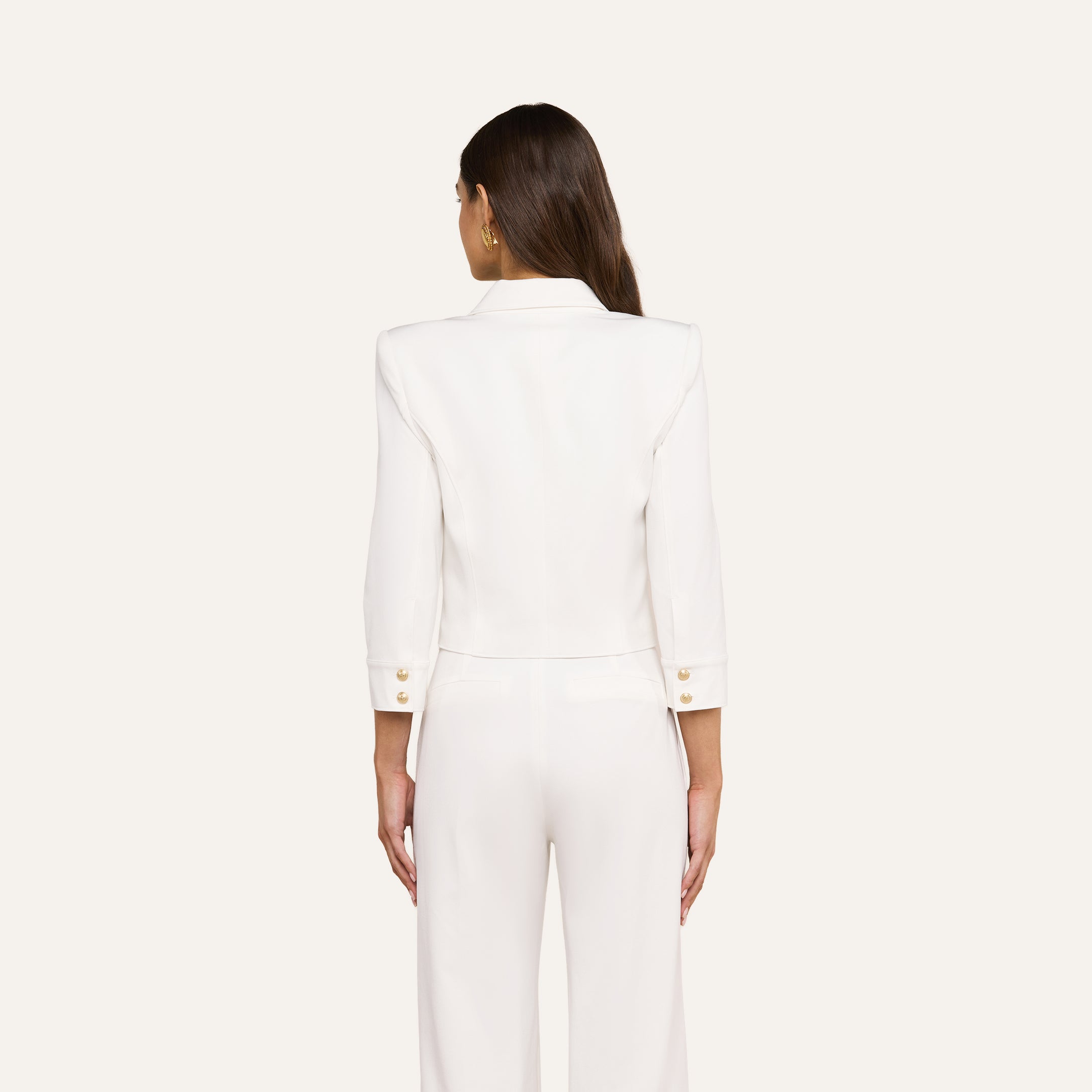 A woman with long brown hair stands facing away, wearing the Kumi Cropped Jacket in white stretch-suiting fabrication with gold embossed dome buttons and matching white pants against a plain light background.