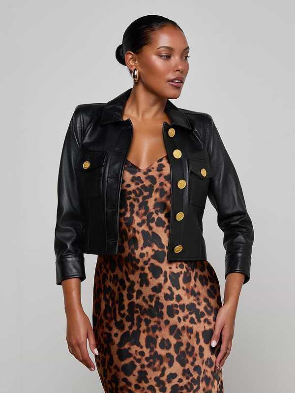 A woman poses against a plain light background, wearing the Kumi Cropped Leather Jacket with gold buttons over a sleeveless leopard print dress. The three-quarter sleeve leather jacket adds an edgy touch to her chic ensemble.