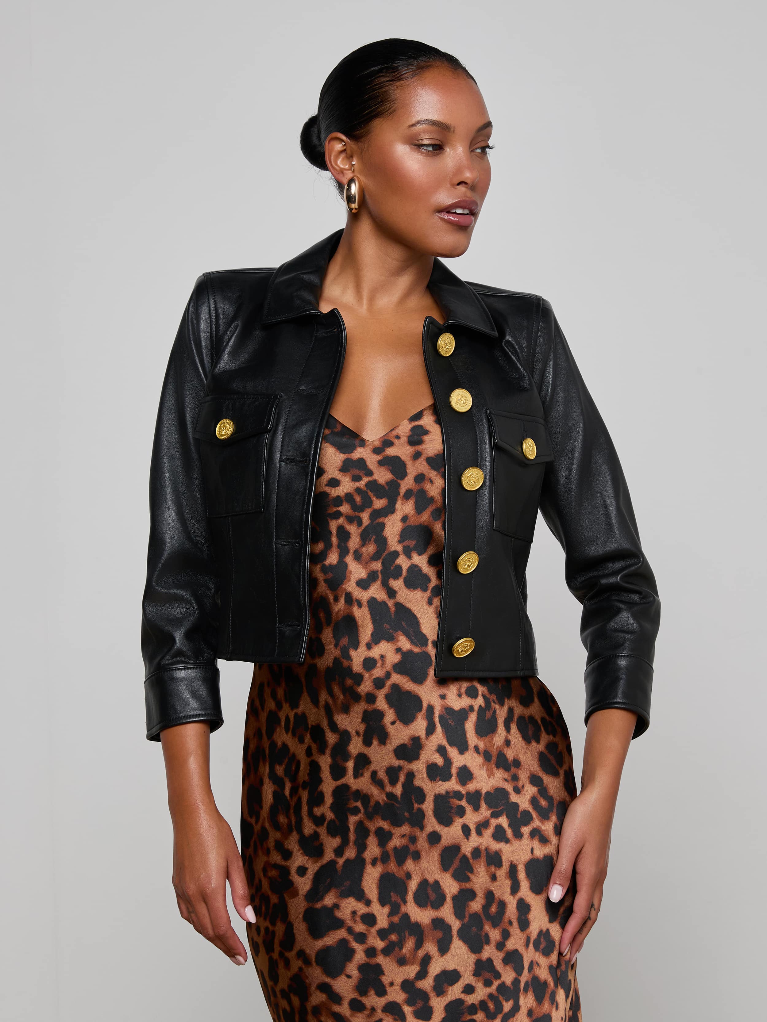 A woman poses against a plain light background, wearing the Kumi Cropped Leather Jacket with gold buttons over a sleeveless leopard print dress. The three-quarter sleeve leather jacket adds an edgy touch to her chic ensemble.