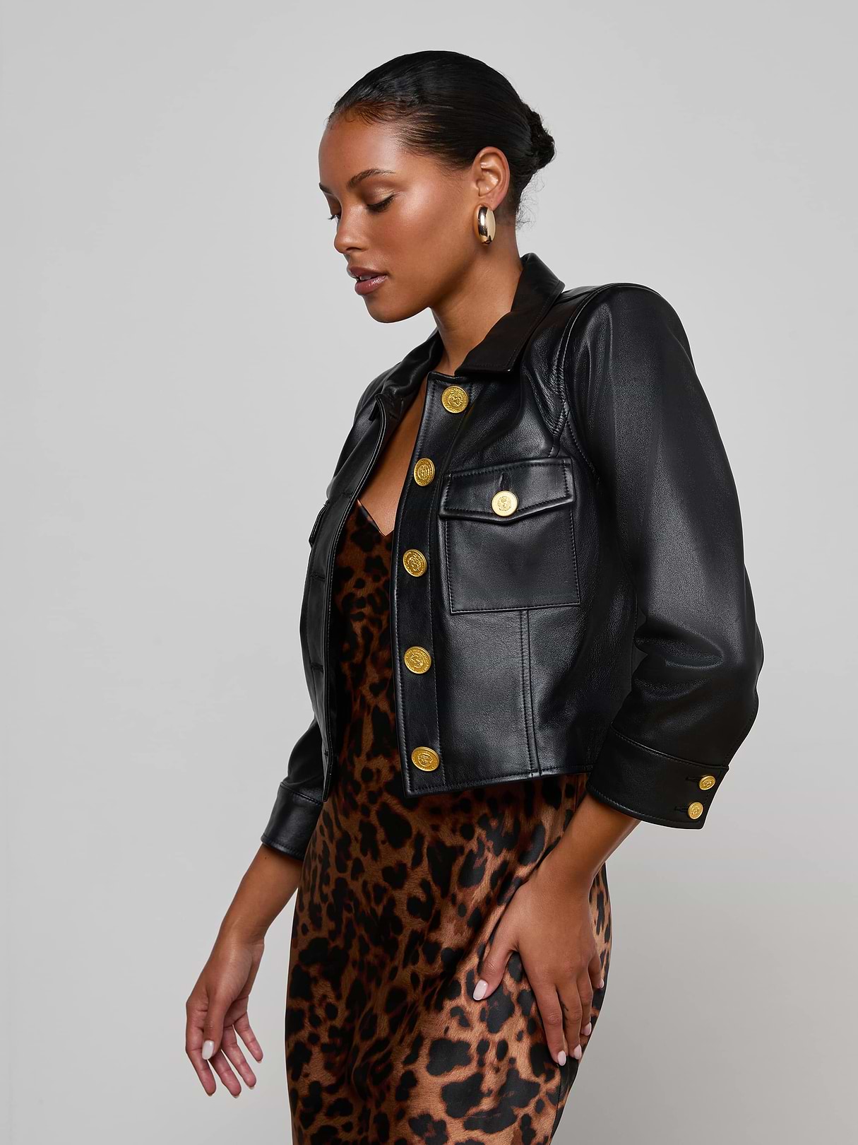 Wearing the Kumi Cropped Leather Jacket—a black lambskin piece with three-quarter sleeves and gold buttons—over a leopard-print dress, a woman stands against a plain light background, looking down to the side.
