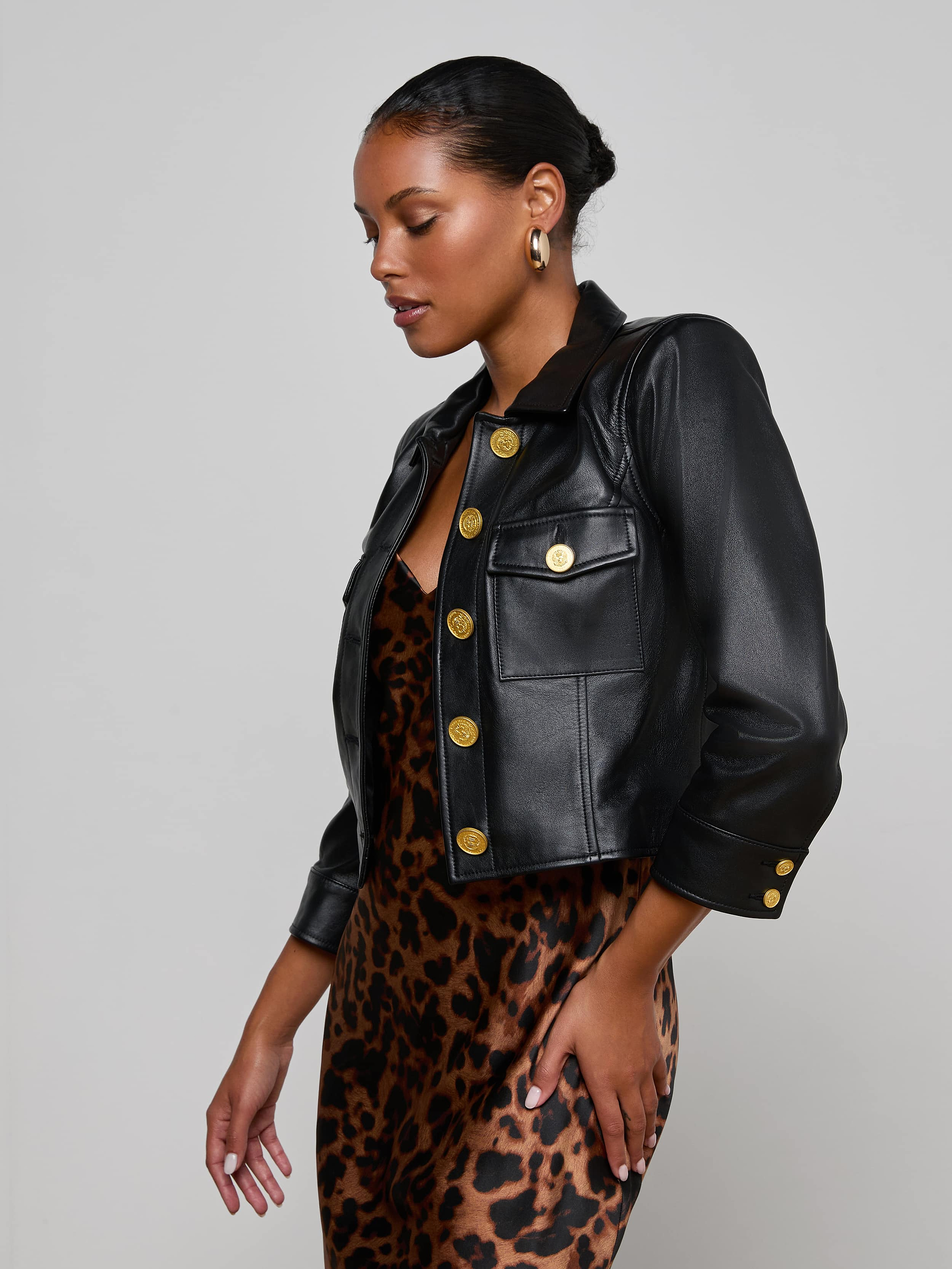 Wearing the Kumi Cropped Leather Jacket—a black lambskin piece with three-quarter sleeves and gold buttons—over a leopard-print dress, a woman stands against a plain light background, looking down to the side.