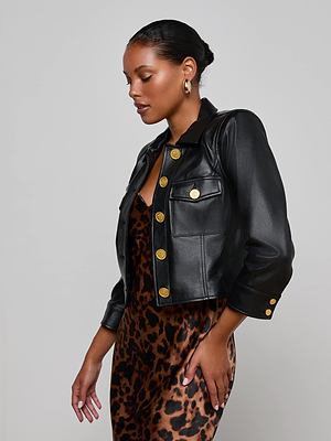 A woman poses in profile against a plain light gray background, wearing the Kumi Cropped Leather Jacket with gold buttons and hoop earrings over a sleeveless leopard print dress.