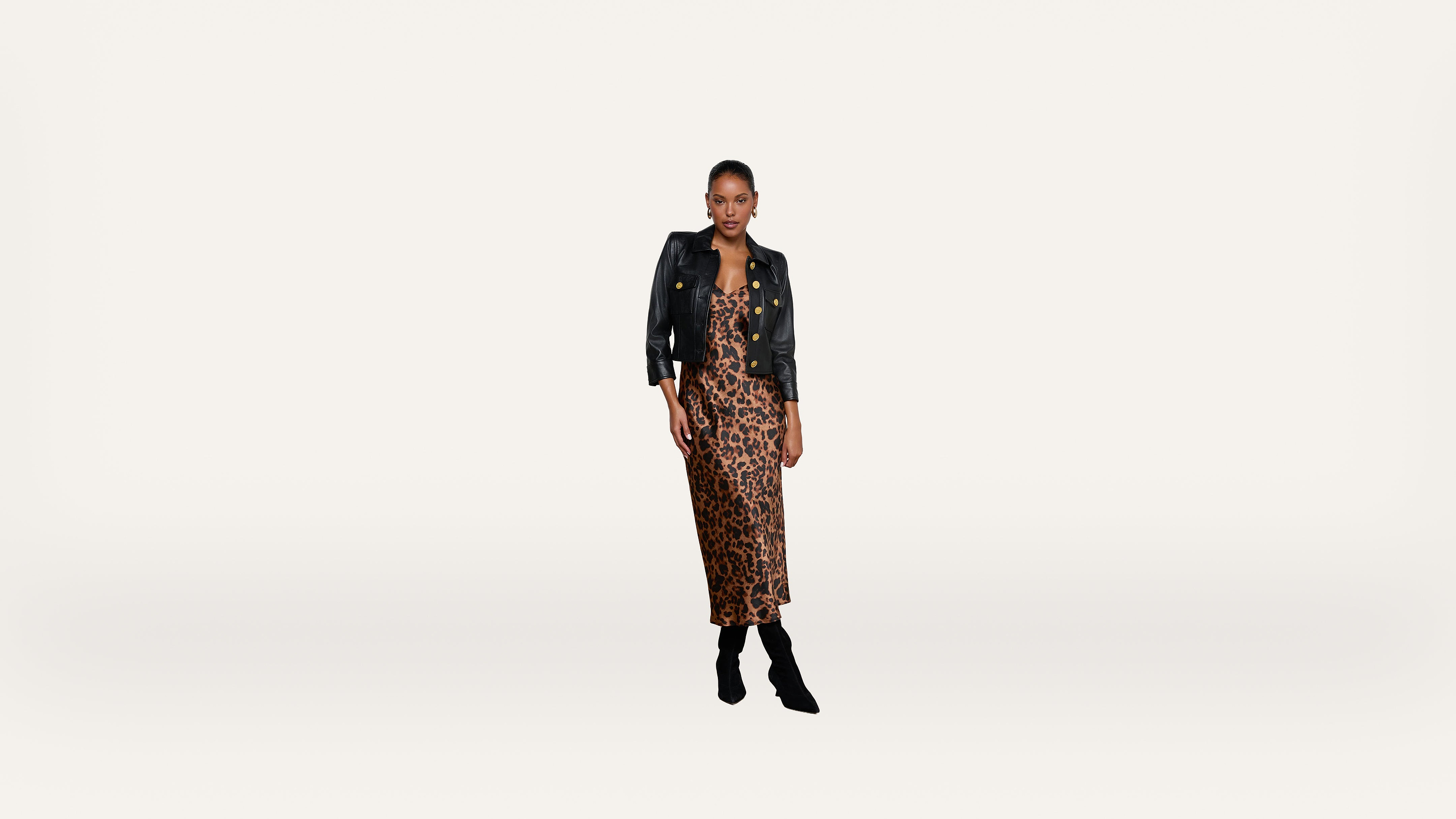 Woman wearing a black leather jacket over a leopard-print dress and black boots stands against a plain white background.