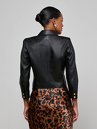 A woman is shown from the back, wearing the Kumi Cropped Leather Jacket in black lambskin with gold buttons, paired with a brown leopard print skirt. Her hair is styled in a low bun and she accessorizes with silver hoop earrings. The background is plain and light-colored.