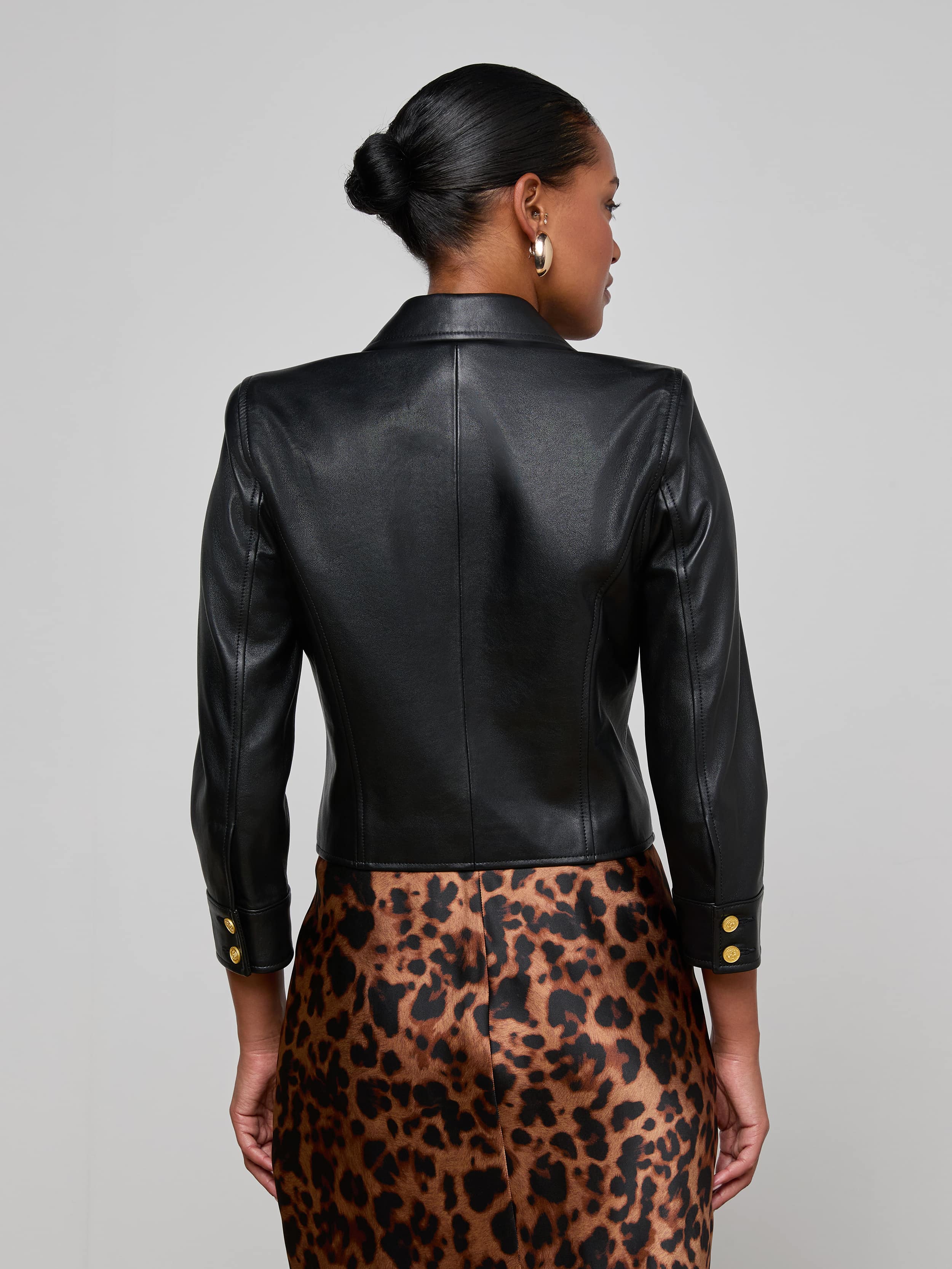A woman is shown from the back, wearing the Kumi Cropped Leather Jacket in black lambskin with gold buttons, paired with a brown leopard print skirt. Her hair is styled in a low bun and she accessorizes with silver hoop earrings. The background is plain and light-colored.