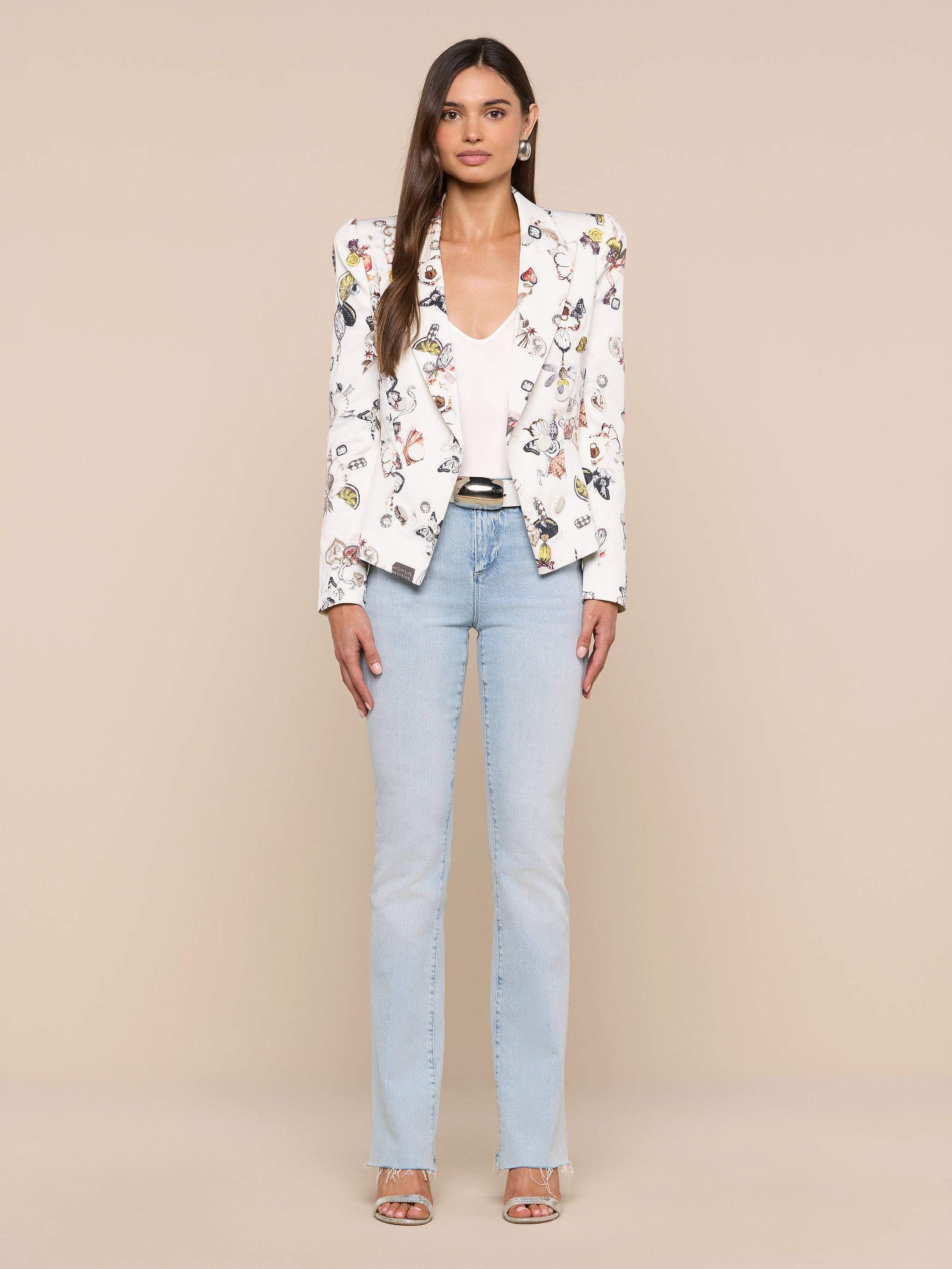 A woman stands facing forward against a plain white background, wearing the Bethany Structured Blazer in a figure-flattering white floral print, styled with a white top, light blue jeans, and open-toe heels.