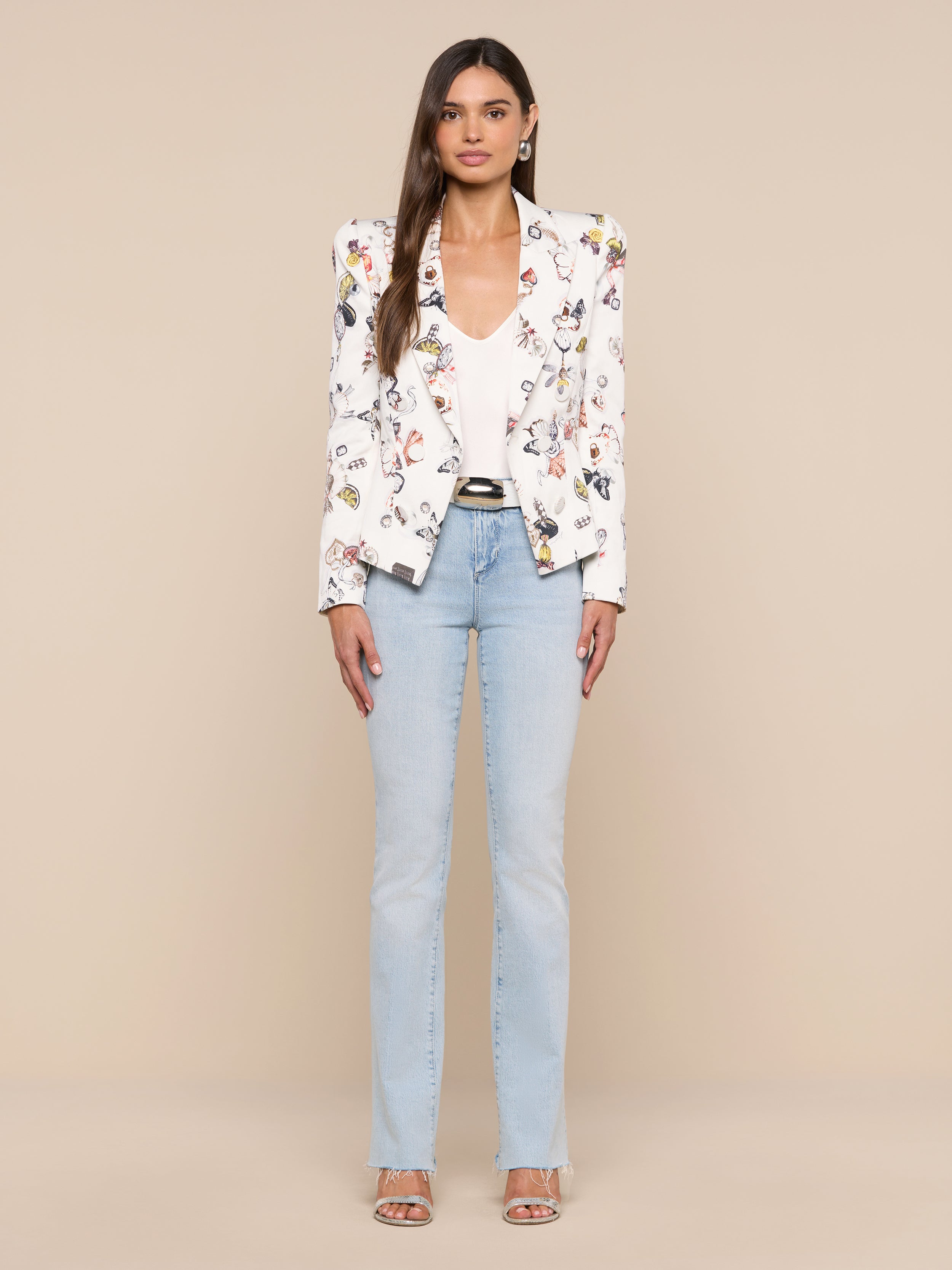 A woman stands facing forward against a plain white background, wearing the Bethany Structured Blazer in a figure-flattering white floral print, styled with a white top, light blue jeans, and open-toe heels.