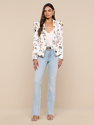 A woman with long brown hair and a neutral expression stands against a plain background, wearing the Bethany Structured Blazer featuring a white floral design, paired with a white top, light blue jeans, silver heels, and earrings.