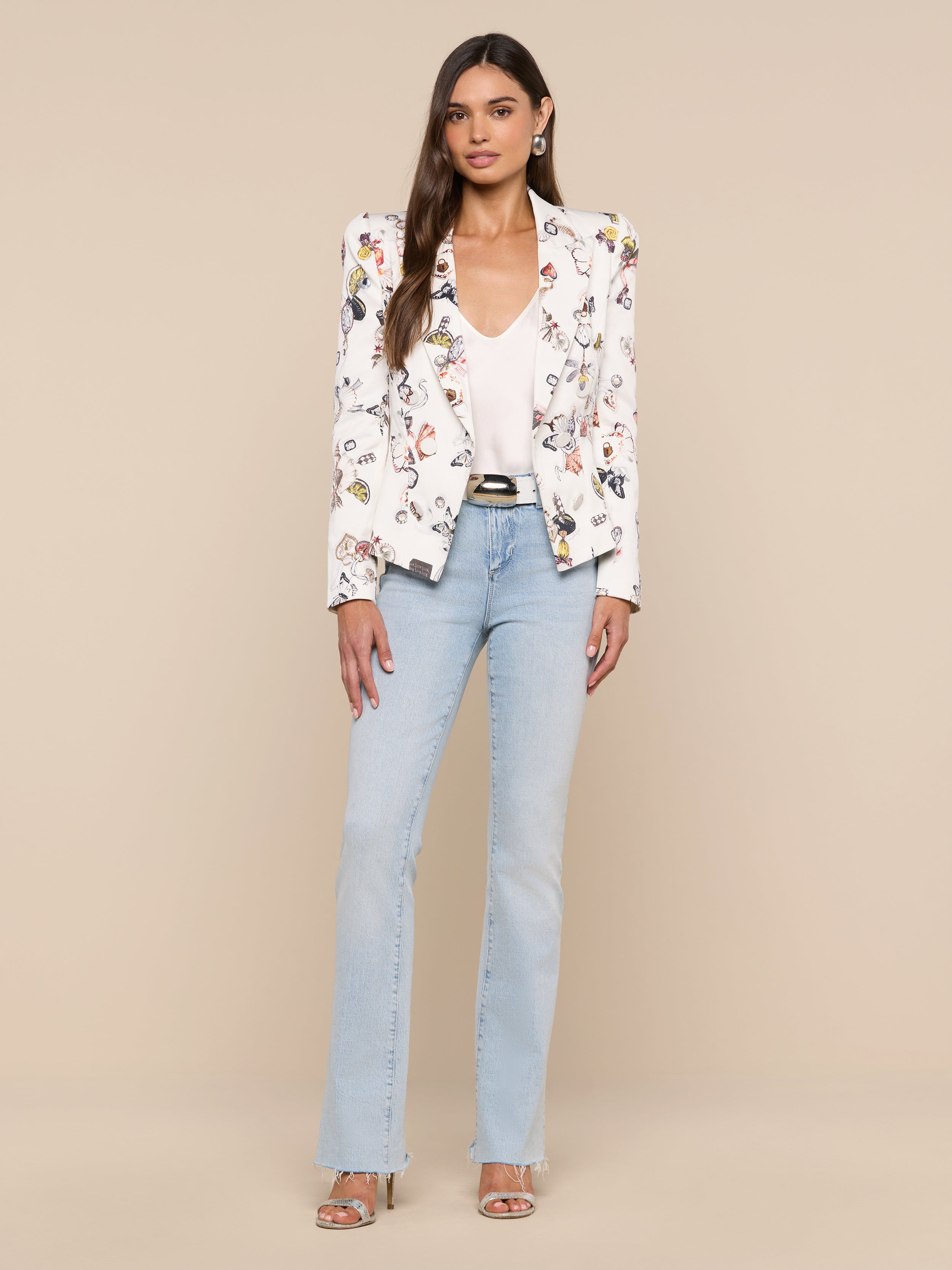 A woman with long brown hair and a neutral expression stands against a plain background, wearing the Bethany Structured Blazer featuring a white floral design, paired with a white top, light blue jeans, silver heels, and earrings.