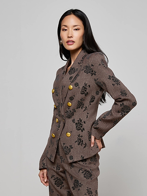 A woman with long dark hair models the L’AGENCE Bethany Flocked Herringbone Blazer, which features a black floral pattern and gold buttons, paired with matching pants, as she poses against a plain light background.