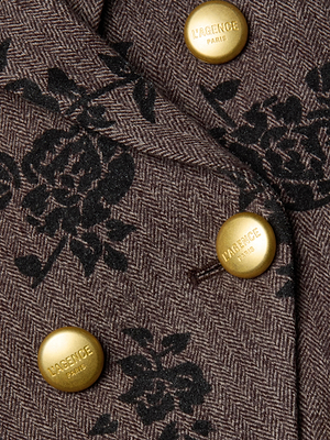 Close-up of the Bethany Flocked Herringbone Blazer by L’AGENCE shows brown herringbone fabric with black floral prints, accented by three gold L’AGENCE PARIS-engraved buttons for a refined touch.