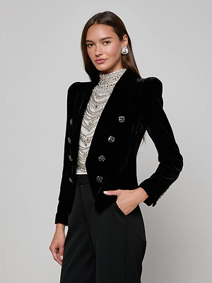 A woman with long brown hair models the L'AGENCE Bethany Velvet Blazer featuring crystal-embellished buttons, a beaded high-neck top, black trousers, and statement earrings, posing with one hand in her pocket against a gray background.