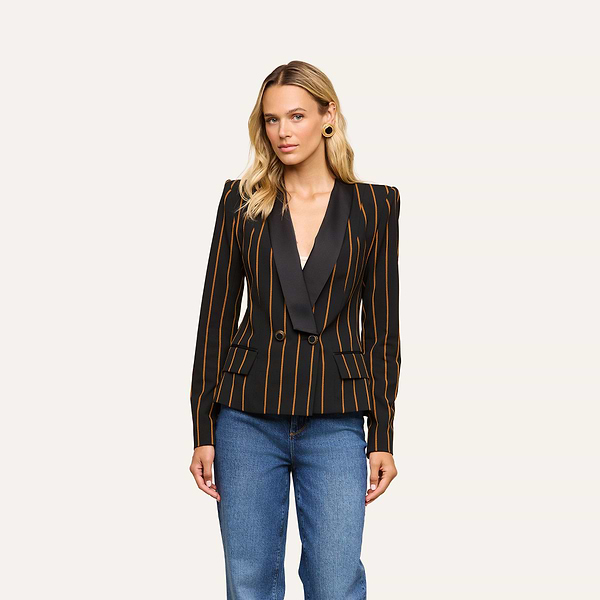 A woman with long blonde hair wears the Imogen Satin Lapel Blazer—a black double-breasted blazer featuring gold vertical stripes and dark satin lapels—paired with blue jeans, standing against a plain white background.