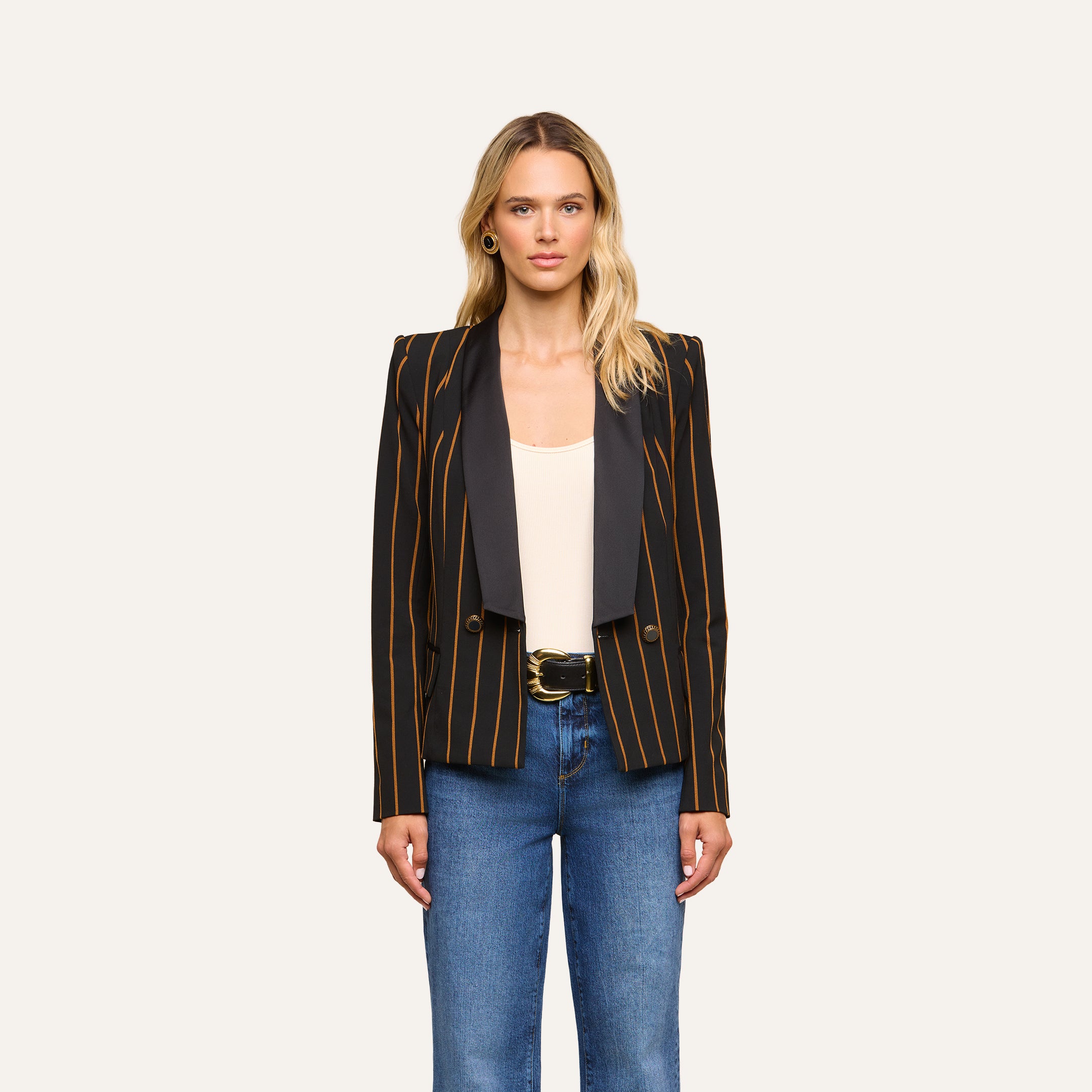 A woman faces forward against a plain light background, wearing the Imogen Satin Lapel Blazer—a double-breasted black blazer with gold vertical stripes and a satin lapel—paired with a white top, blue jeans, and a black belt with a gold buckle.