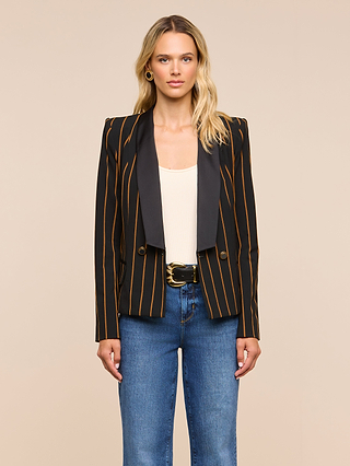 A woman with long blonde hair and a neutral expression stands against a beige background, wearing the Imogen Satin Lapel Blazer—featuring black fabric with orange vertical stripes and a sleek satin lapel—styled over a white top, blue jeans, and finished with a black belt featuring a gold buckle.