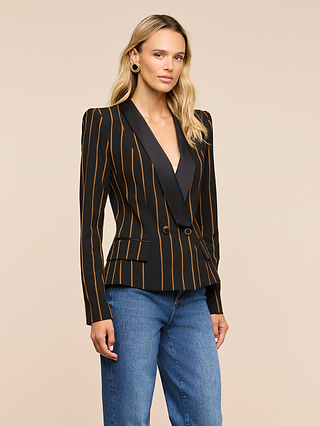 A woman wearing the Imogen Satin Lapel Blazer—a double-breasted black blazer with orange vertical stripes, black satin lapels, and gold buttons—paired with blue jeans, stands against a plain beige background.