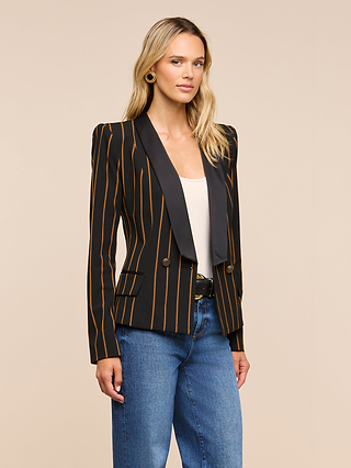A woman with long blonde hair and gold hoop earrings stands against a plain beige background, wearing the Imogen Satin Lapel Blazer—a black double-breasted blazer with orange stripes and a satin lapel—paired with a white top and blue jeans.