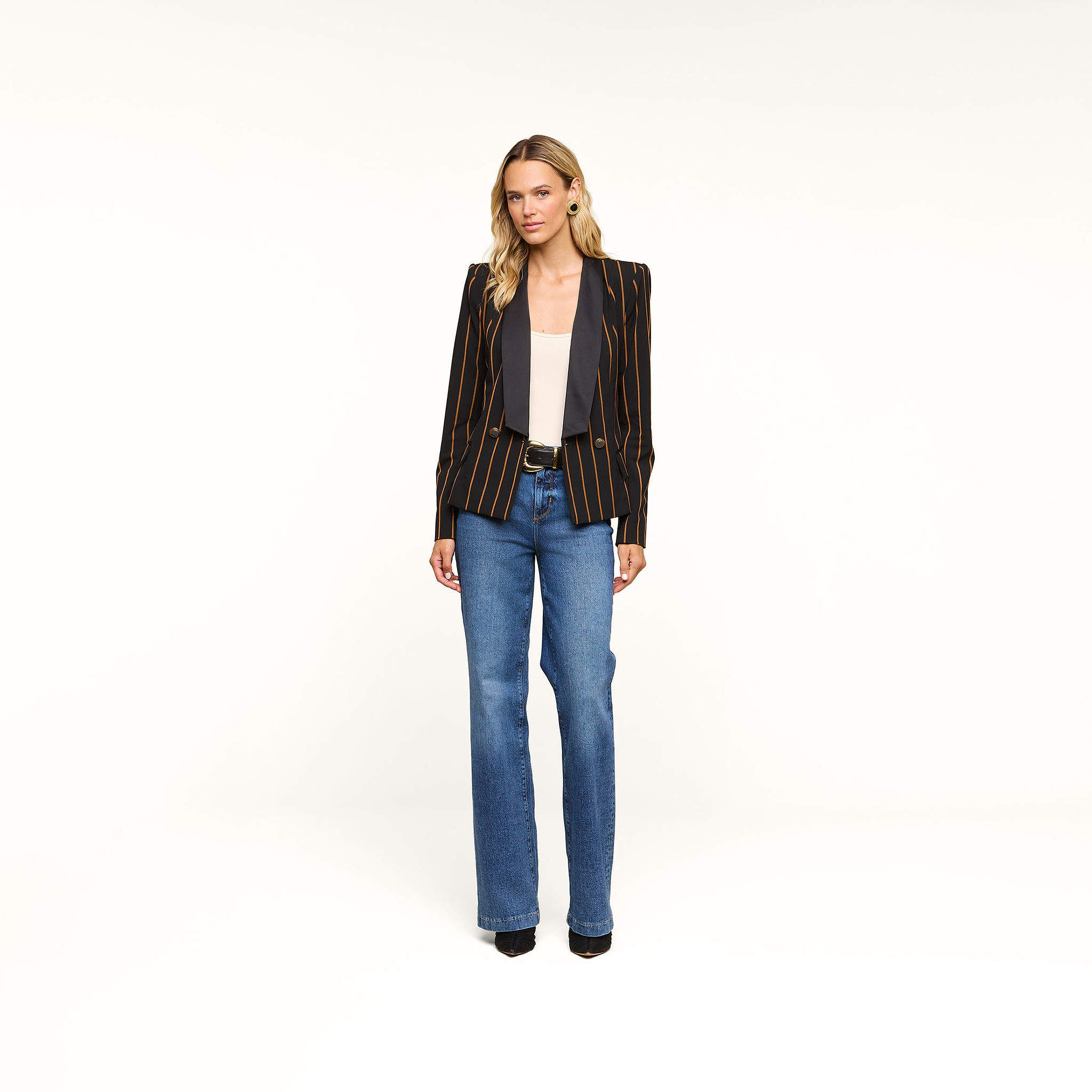 A woman stands against a plain white background, wearing the Imogen Satin Lapel Blazer—a double-breasted blazer with orange stripes and a satin lapel—paired with a gray top, blue jeans, and black heels.