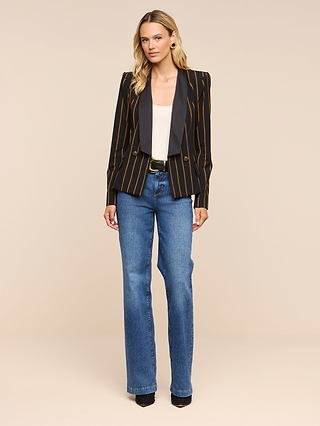 A woman stands against a beige background, wearing the Imogen Satin Lapel Blazer—a black double-breasted blazer with orange stripes and a satin lapel—paired with a white top, blue jeans, a black belt, and black shoes. She has blonde hair and looks forward with a neutral expression.