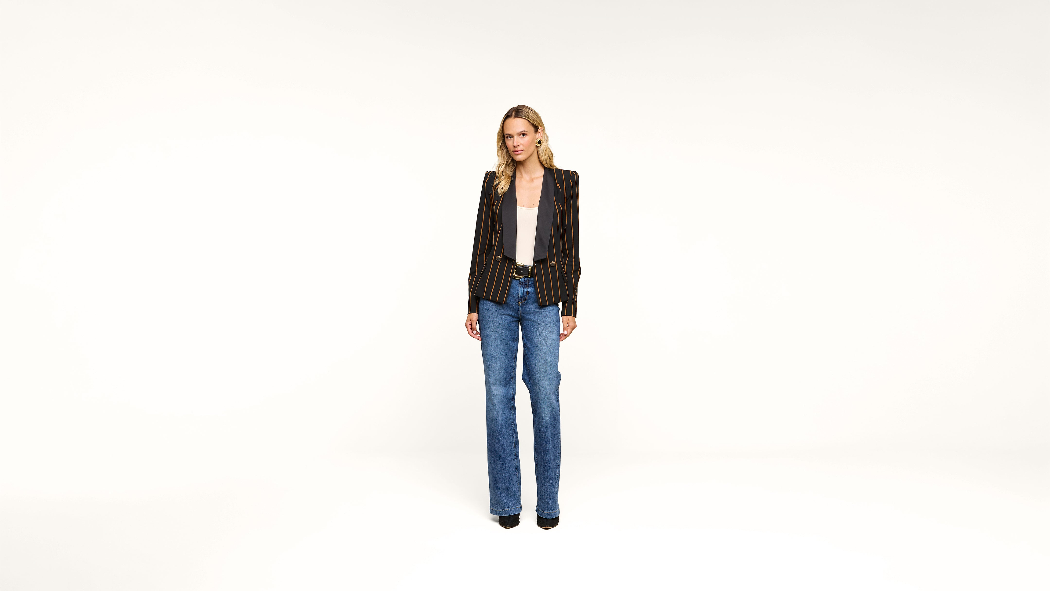 A woman stands against a plain white background, wearing a black striped blazer over a white top, blue jeans, and black shoes. She has long hair and a neutral expression.